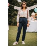 Mladá žena stojí venku na půlroční oslavě v pastelové blůze a kalhotách, s jemnými šperky a teniskami.