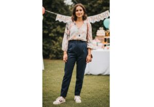 Mladá žena stojí venku na půlroční oslavě v pastelové blůze a kalhotách, s jemnými šperky a teniskami.