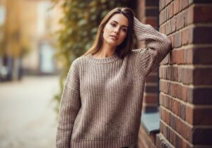 Mladá žena v oversized pleteném svetru opírající se o cihlovou zeď s podzimním pozadím