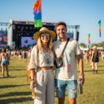Mladý pár na letním hudebním festivalu drží lahve s vodou, stojí na trávě před pódii a vlajkami.